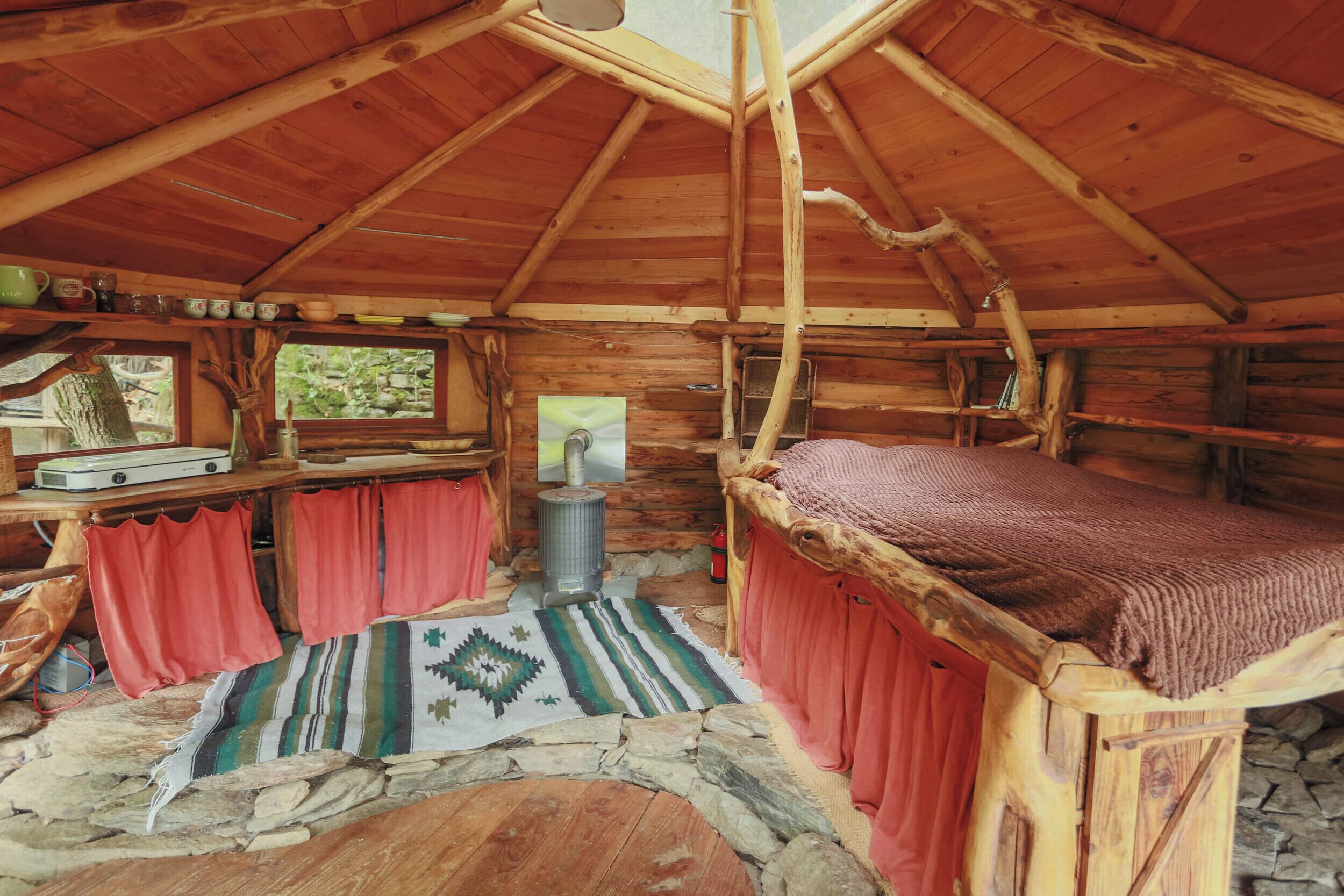 Une cabane en bois rustique, avec un lit, une table et une bouilloire, et une fenêtre donnant sur la verdure.