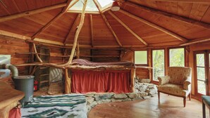 Ein rustikales Blockhäuschen im Inneren mit Holzbalkendecke, einem Bett mit roter Decke, einem Kamin aus Stein und einem Sessel.