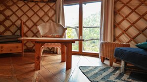 A cozy interior with a wooden table, wicker chairs, a sofa, and a large window offering a view of greenery. - Cévennes yurt (Malons-et-Elze)