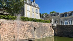 Un mur de pierre ancien avec une fontaine, un grand bâtiment doté de nombreuses fenêtres et un ciel dégagé.