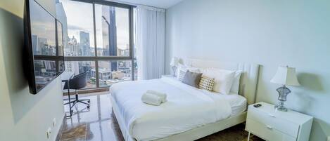 A modern hotel room with a large bed, a desk, a chair, and a view of the city skyline through large windows.