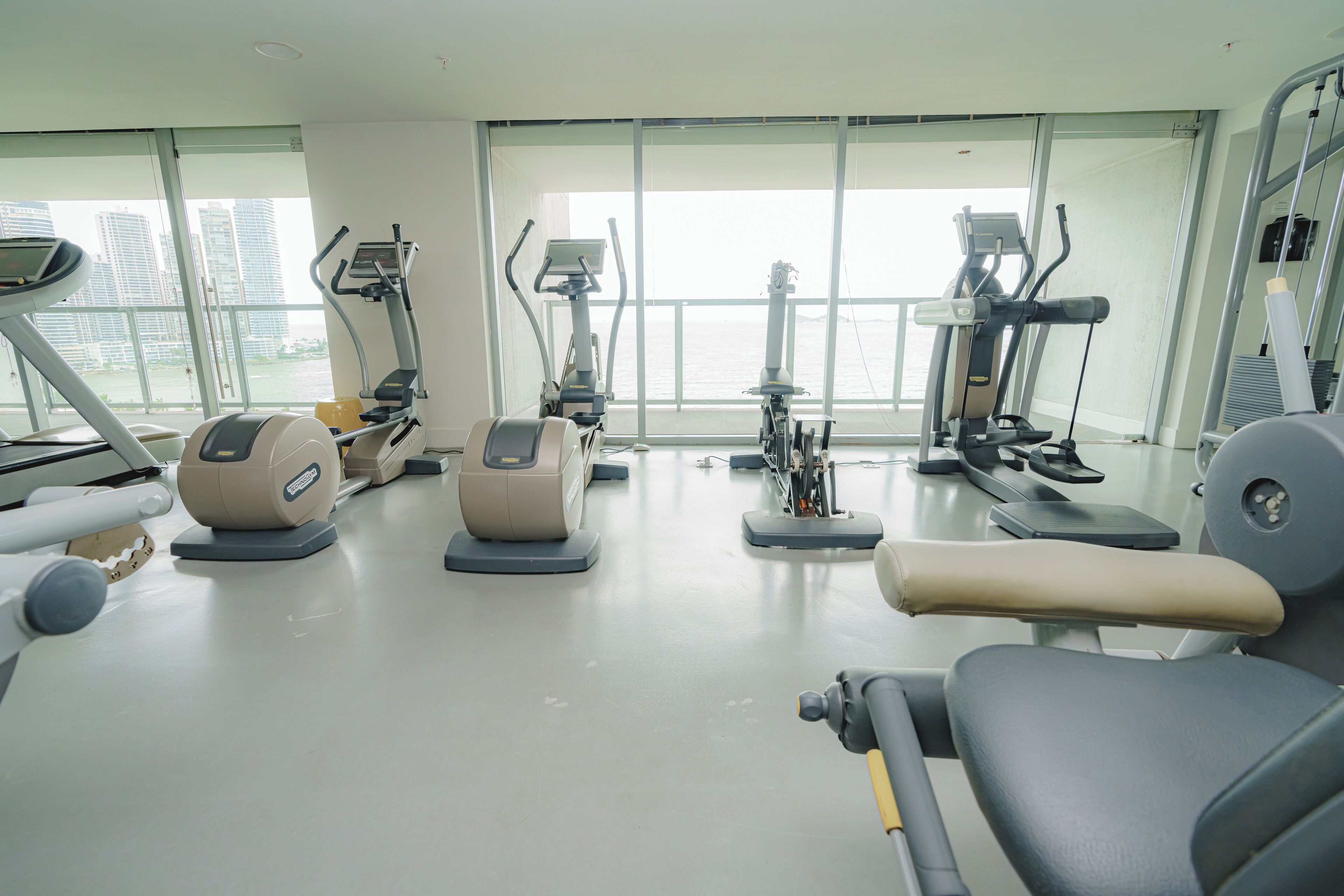 A modern gym with various exercise machines, large windows, and a view of the cityscape.