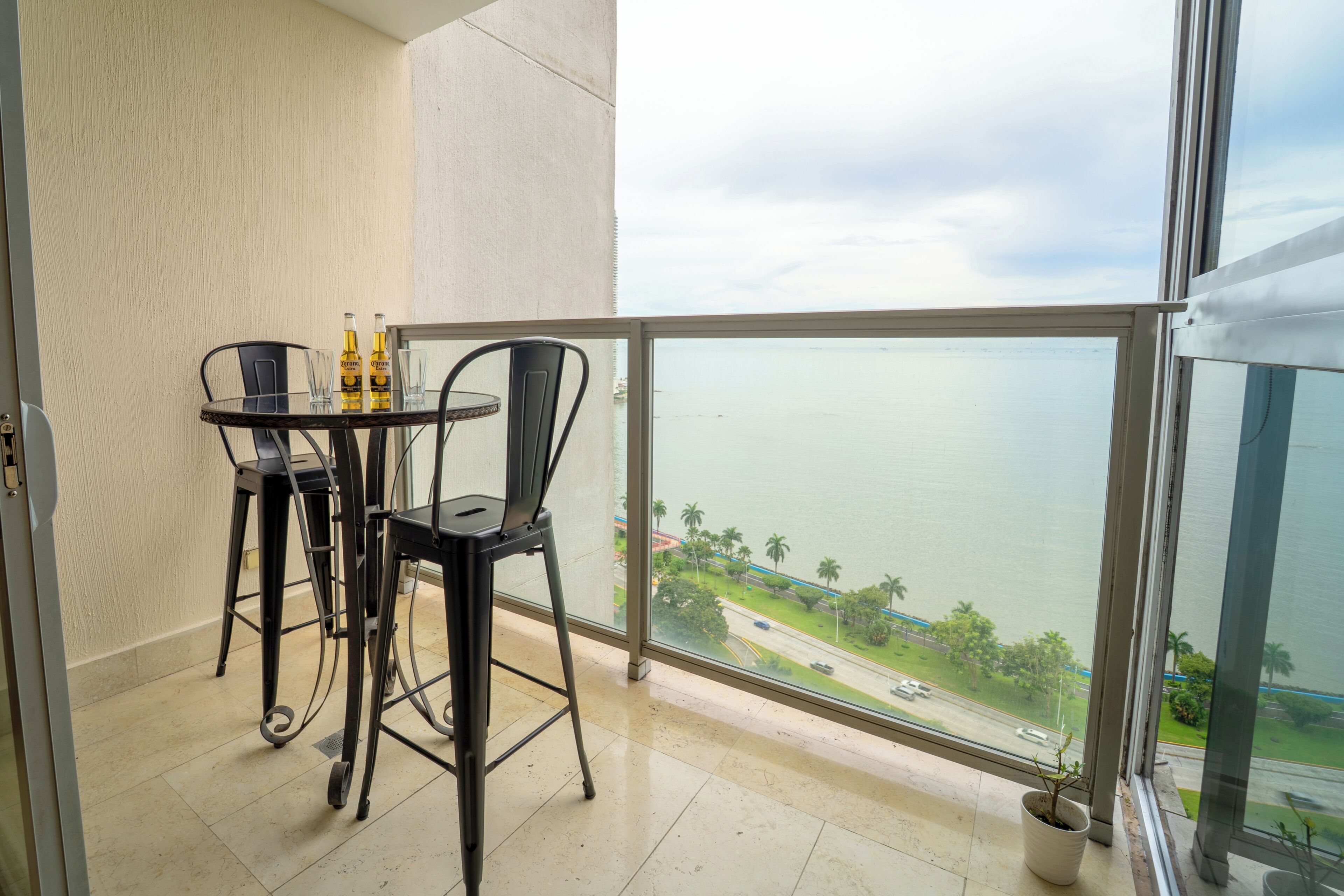 A balcony with a table and chairs, a potted plant, and a view of the sea and a road.