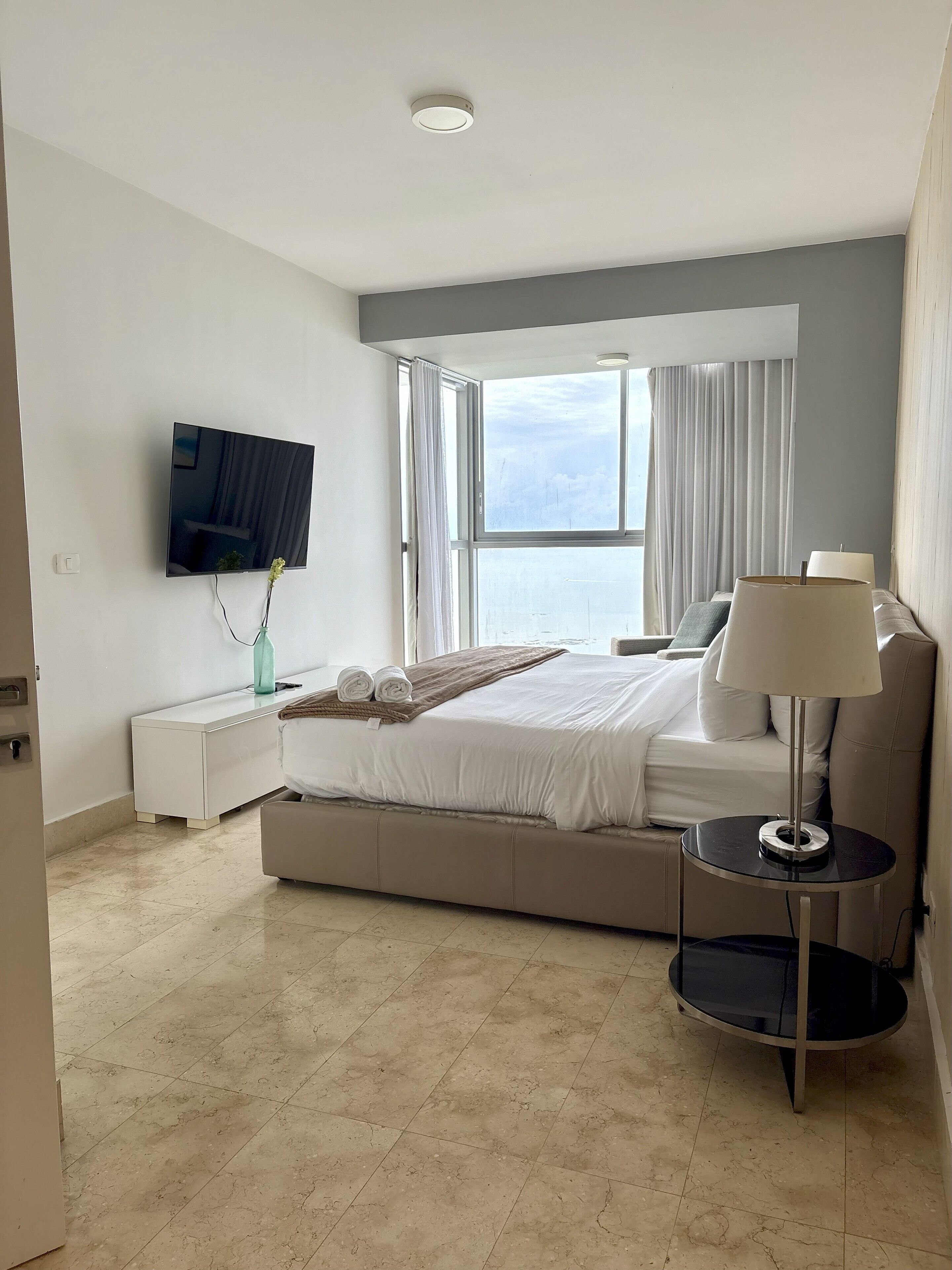 Un hotel moderno con una cama grande, una mesita de noche, un televisor montado en la pared, una ventana con cortinas y vistas a la ciudad.