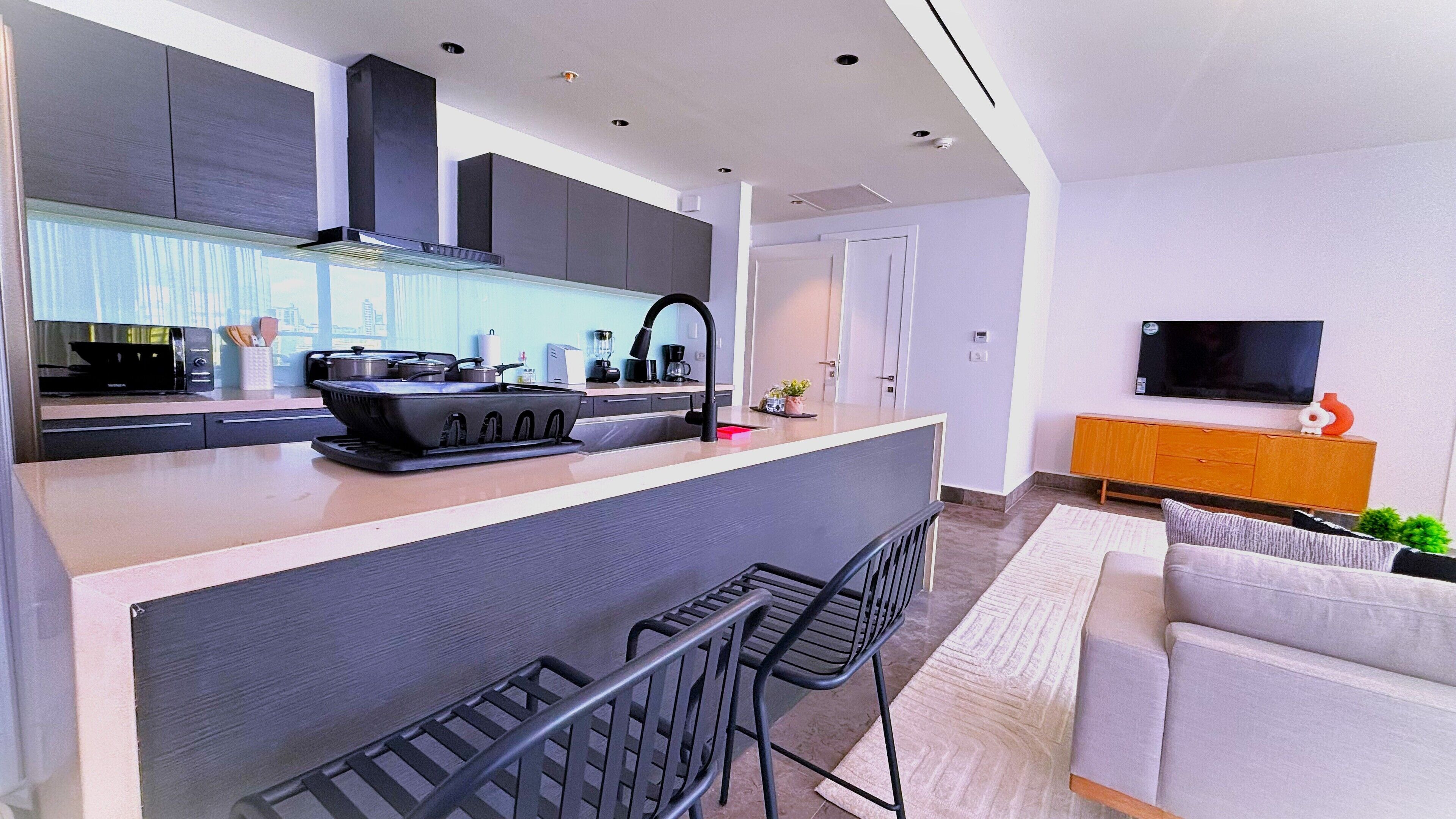 A modern kitchen with a central island, bar stools, a built-in oven, and a flat-screen TV mounted on the wall.