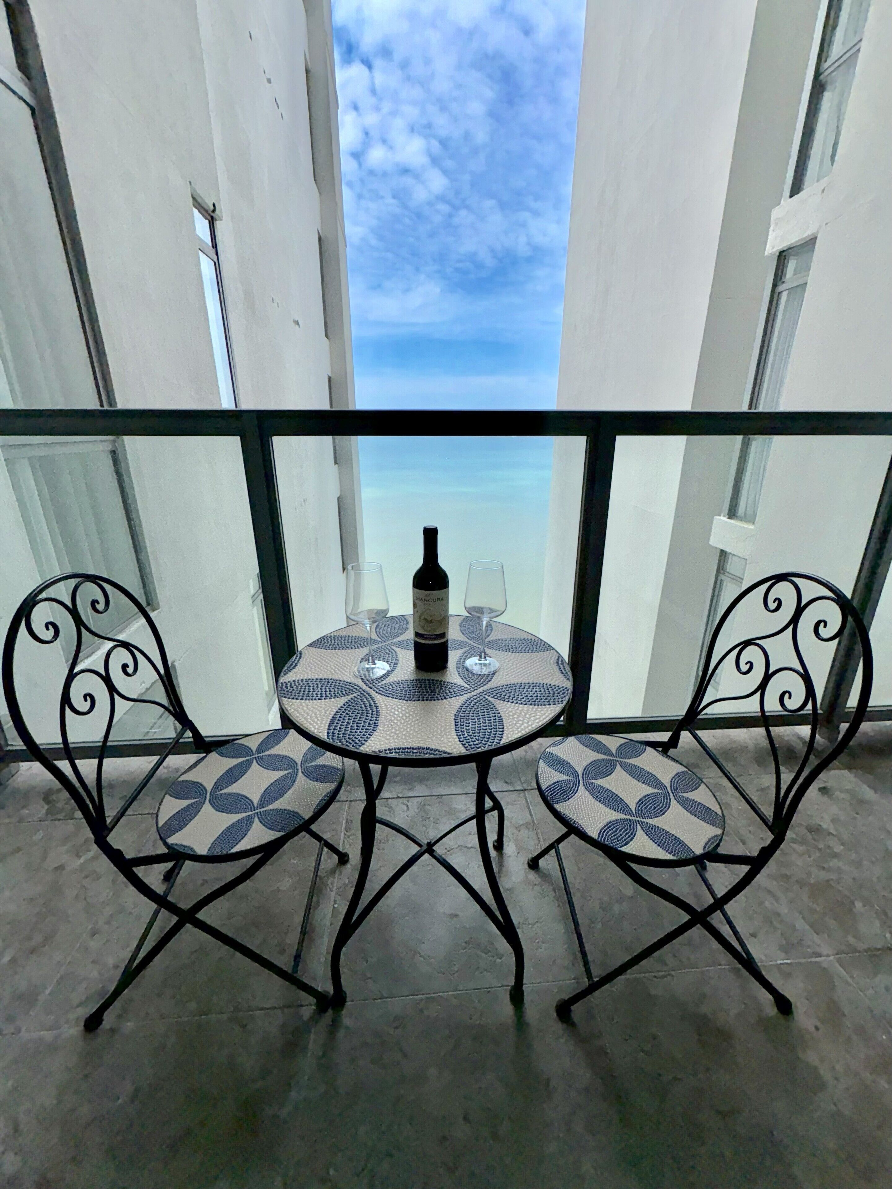 A balcony with a glass railing, two chairs, a round table, and a bottle of wine.