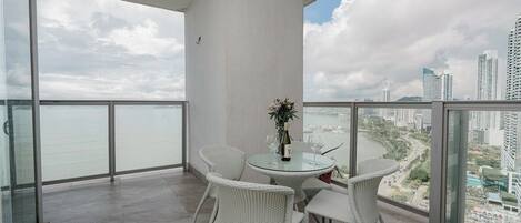 A balcony with a glass table and wicker chairs, overlooking a cityscape and water.