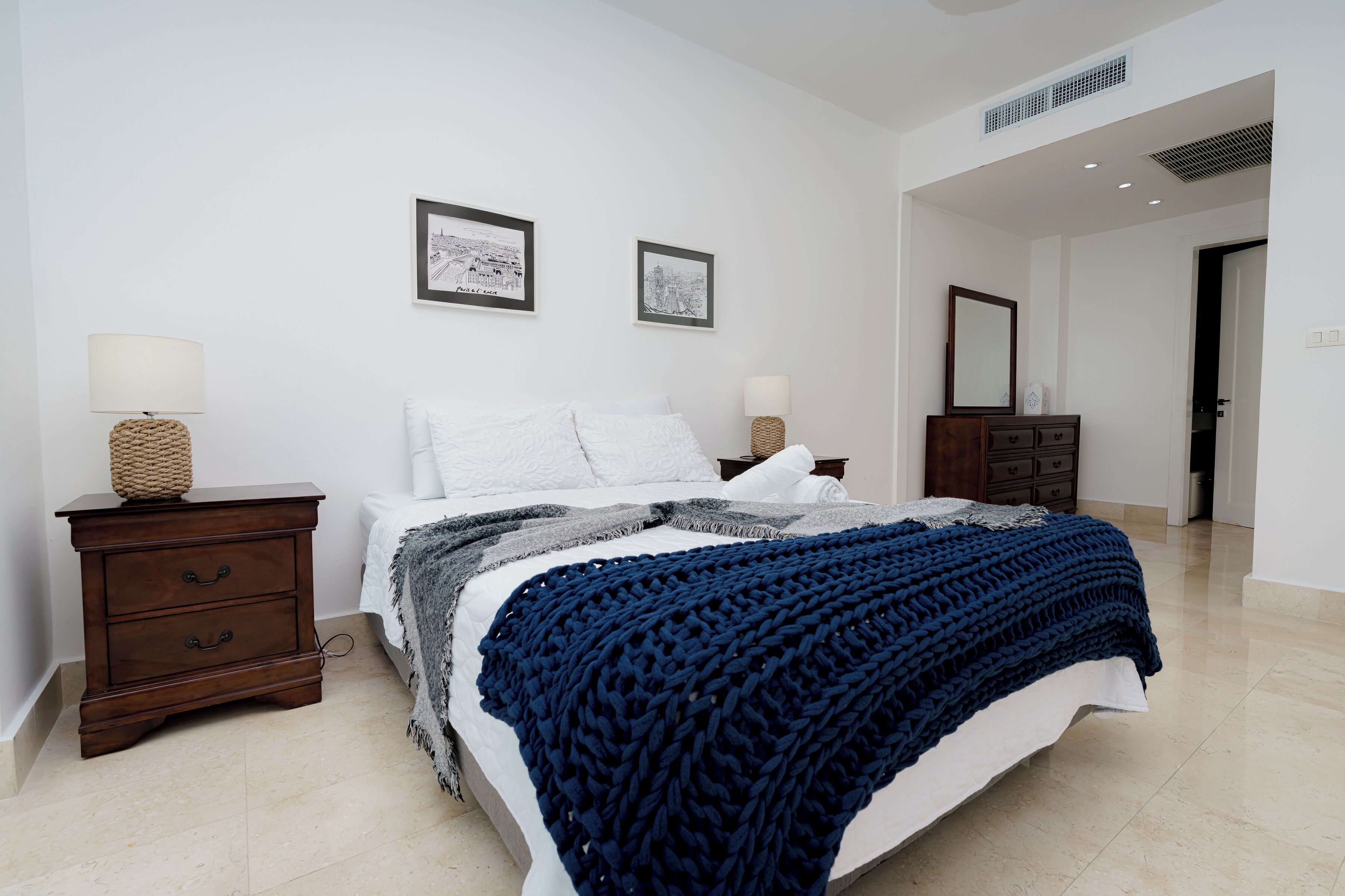 A bedroom with a bed, two bedside tables with lamps, a dresser, and two framed pictures on the wall.