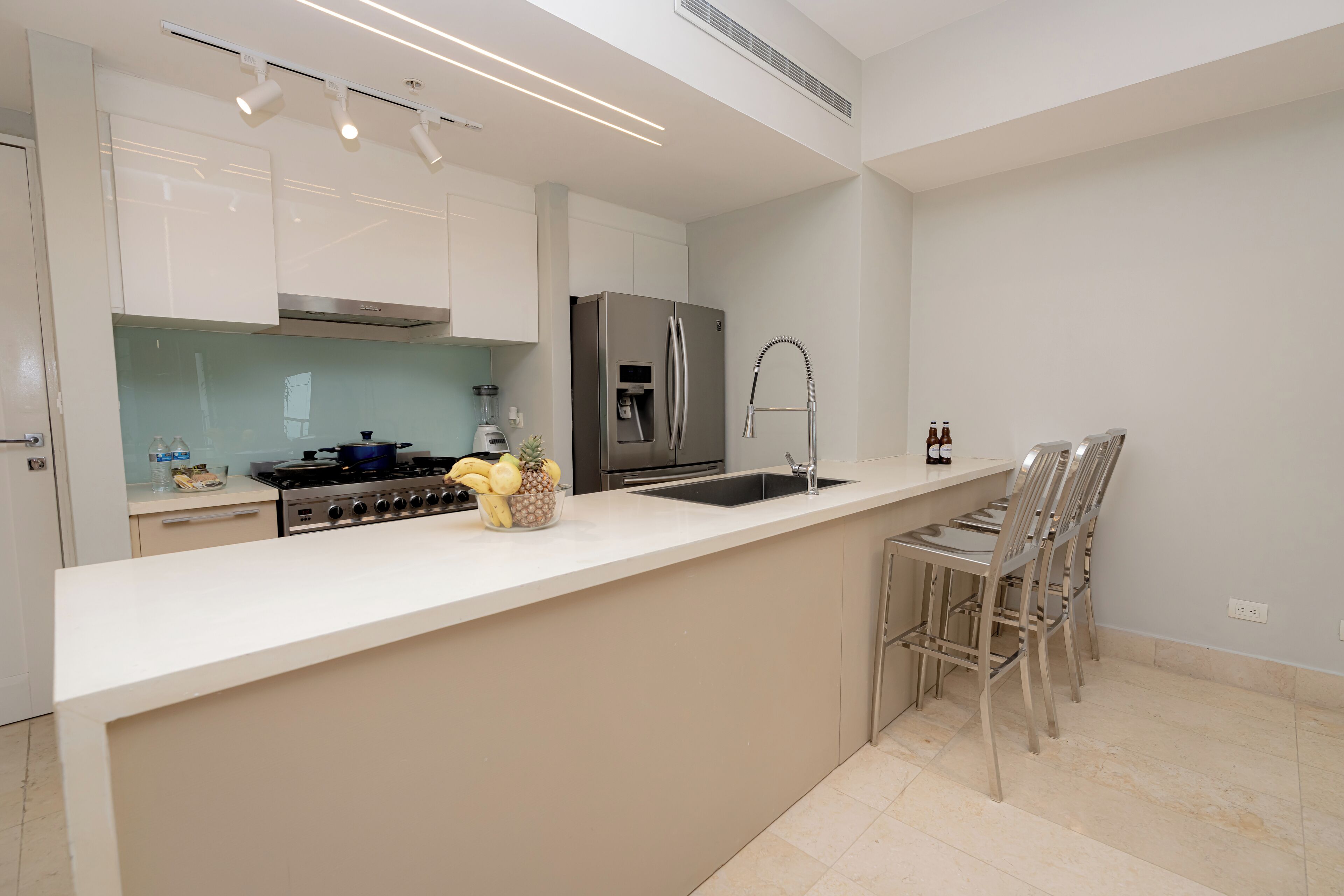 A modern kitchen with white cabinets, a stainless steel refrigerator, a built-in oven, and a kitchen island with chairs.