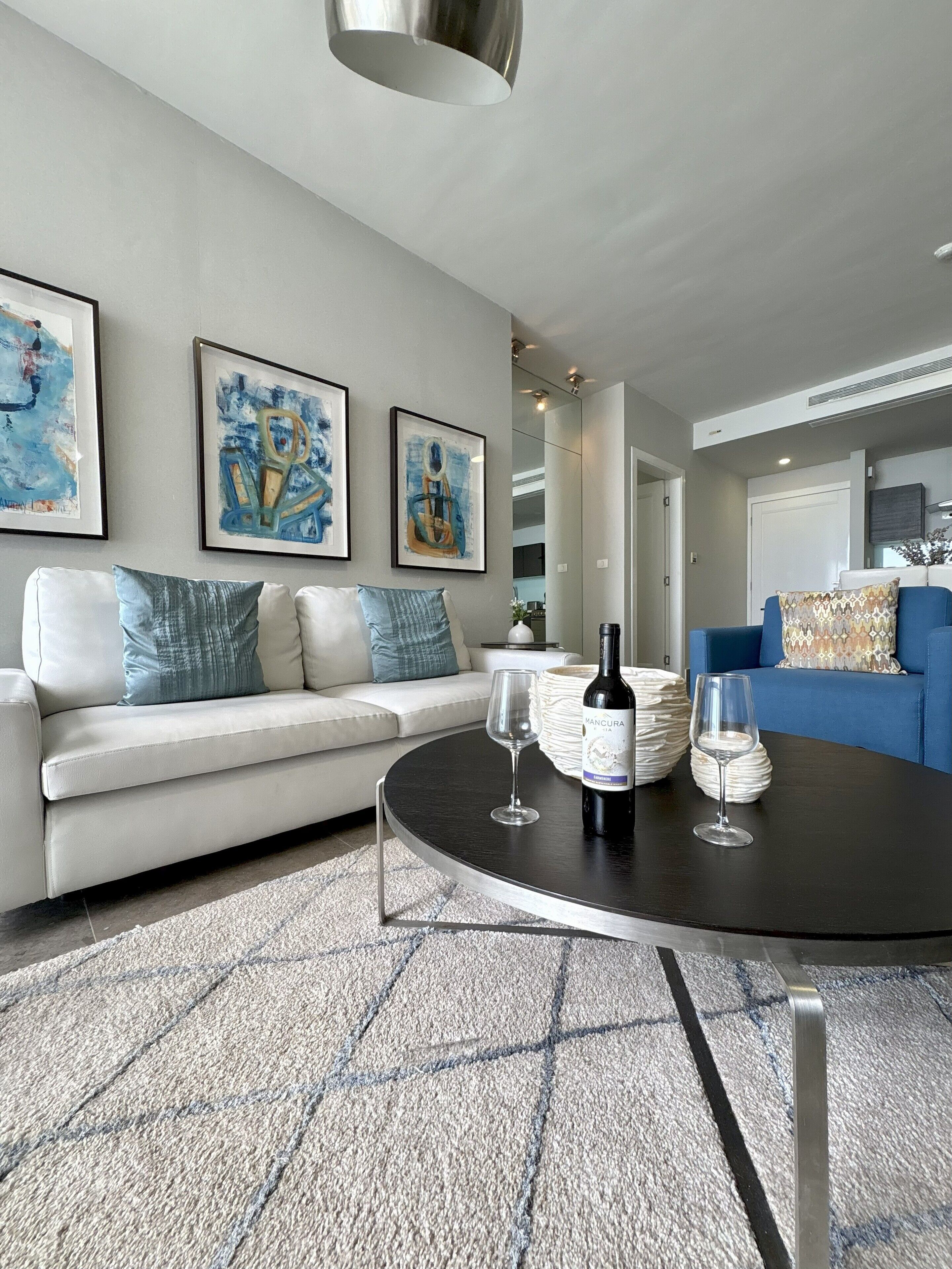 A modern living room with a white sofa, a blue sofa, and a round coffee table with wine glasses and a bottle. There are three framed abstract paintings on the wall.