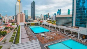 Un espace piscine sur le toit, comprenant plusieurs piscines et des chaises longues, entouré d’immeubles de grande hauteur.