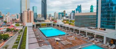A rooftop pool area with multiple swimming pools and lounge chairs, surrounded by high-rise buildings.