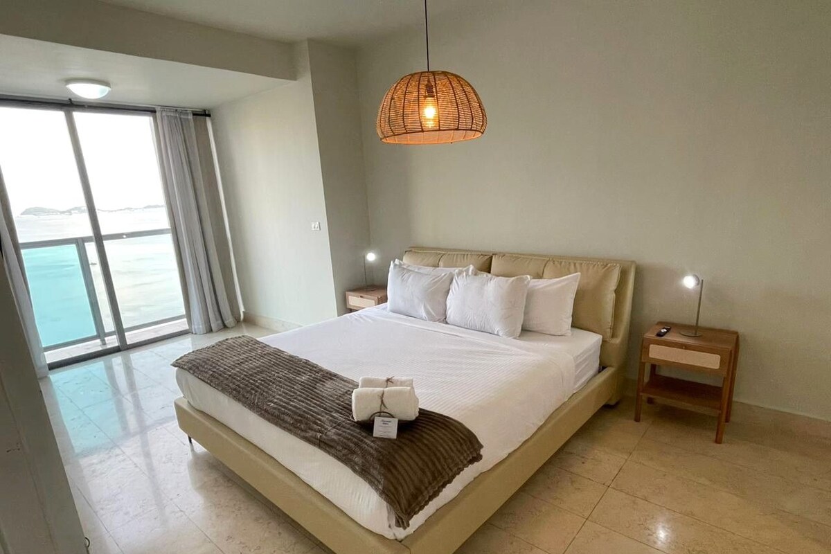 A hotel room with a large bed, a bedside table with a lamp, a view of the sea through a large window, and a hanging light fixture.
