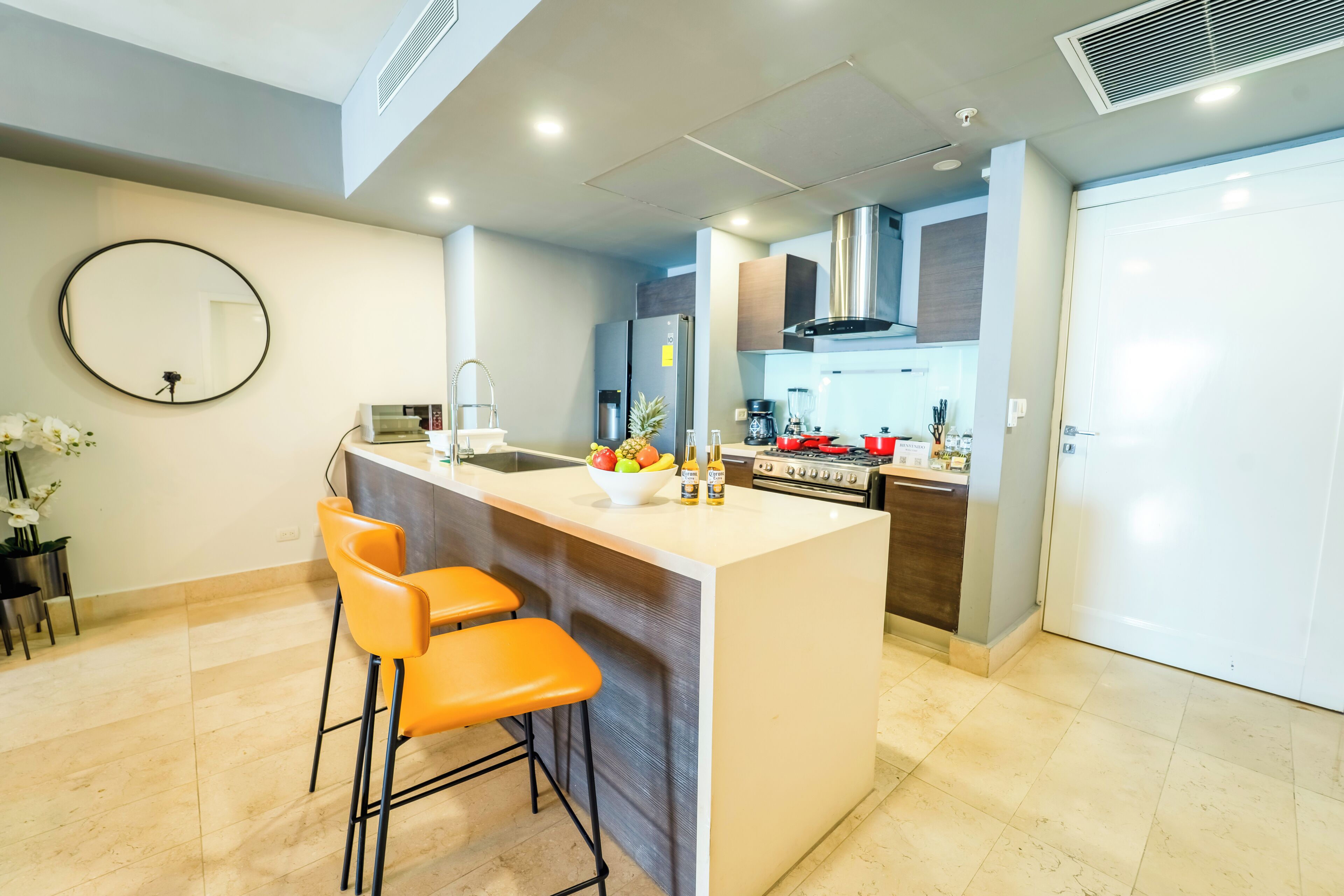 A modern kitchen with a central island, bar stools, a round mirror, and a stainless steel refrigerator.
