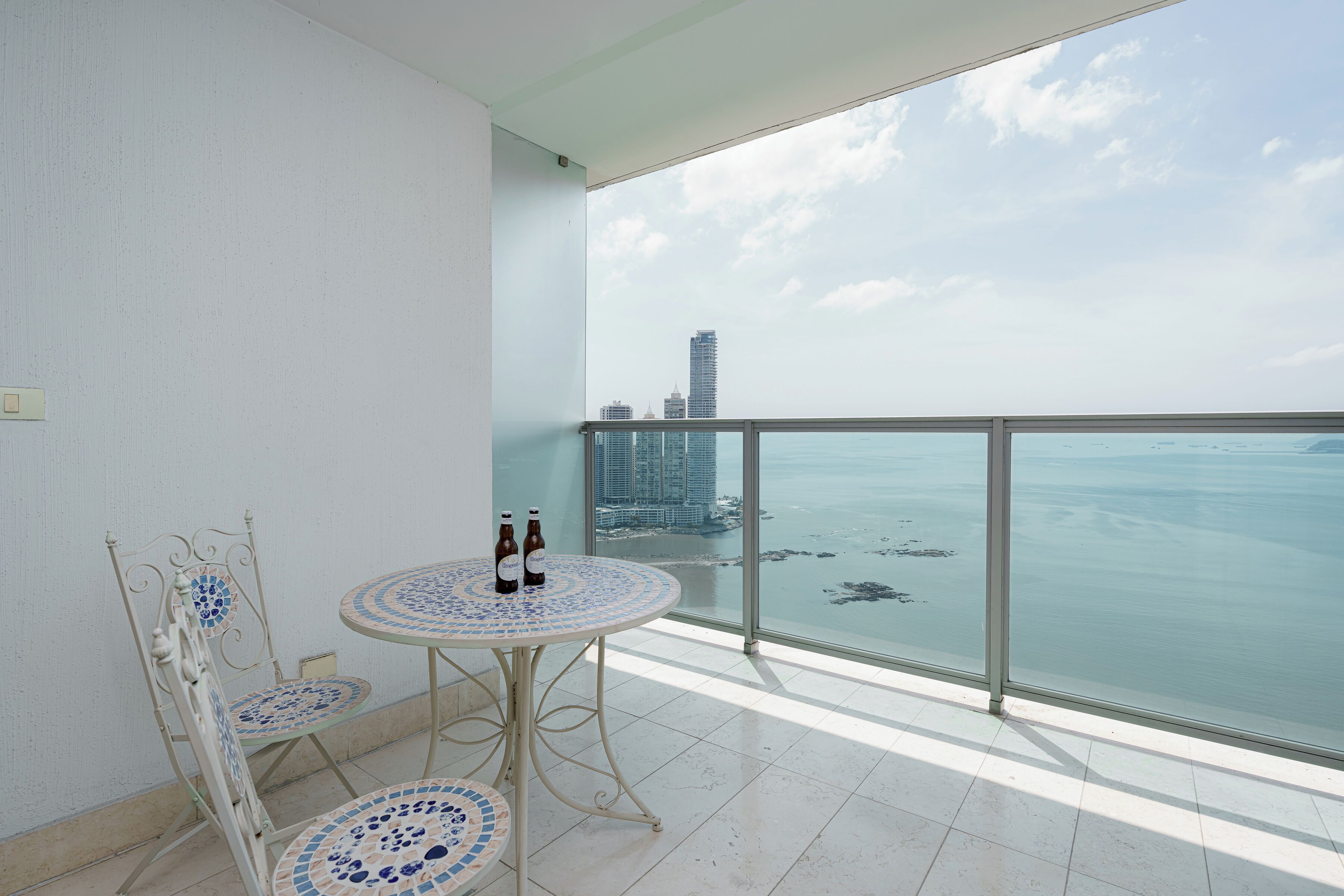 Un balcón con piso de baldosas, dos sillas, una mesita con dos botellas y vistas al mar y edificios.