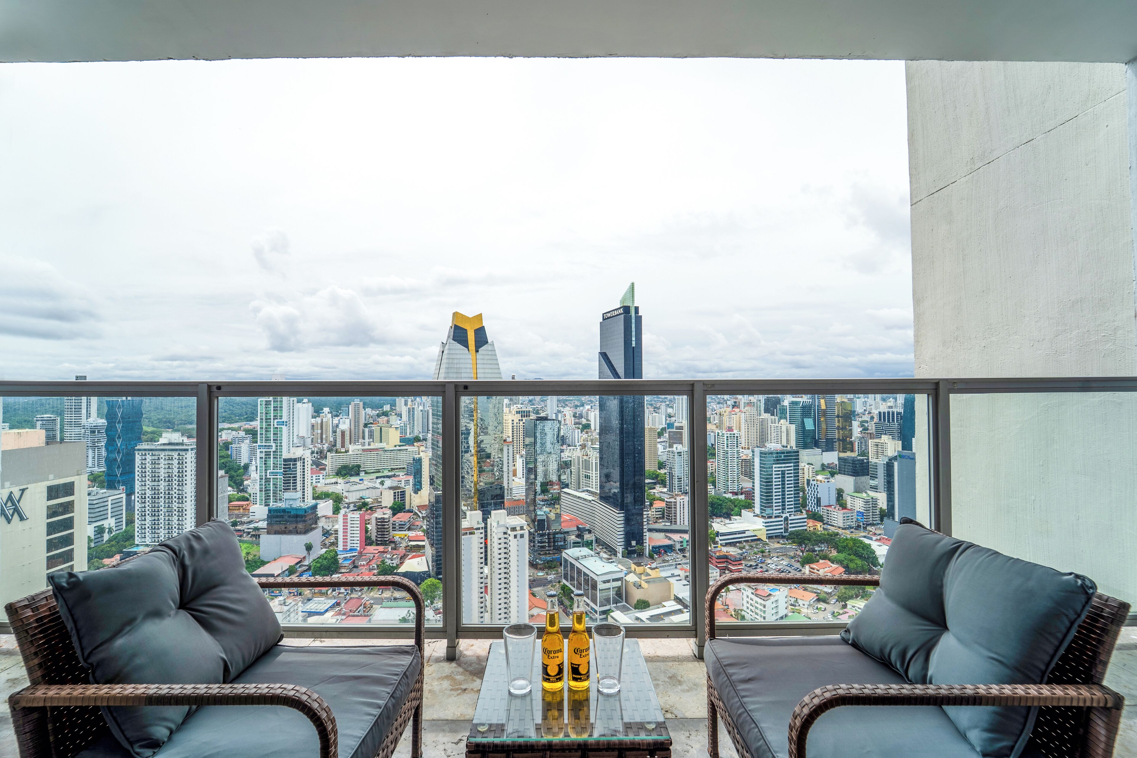 A balcony with wicker chairs and a table, overlooking a cityscape with high-rise buildings.