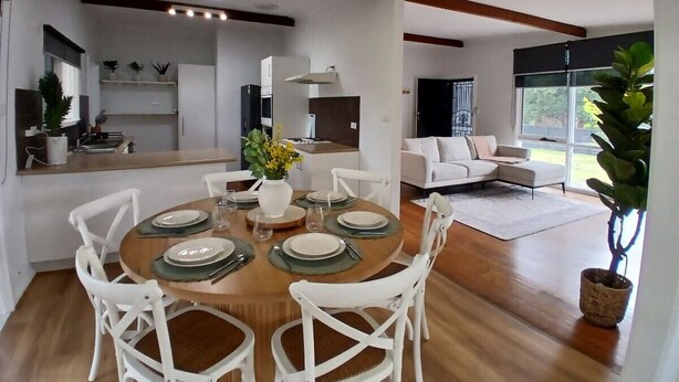 A modern dining area with a wooden table set for six, surrounded by white chairs. A kitchen is visible in the background, and a living area with a sofa and a large window is also visible. - Capel Sound Hideaway (Capel Sound)