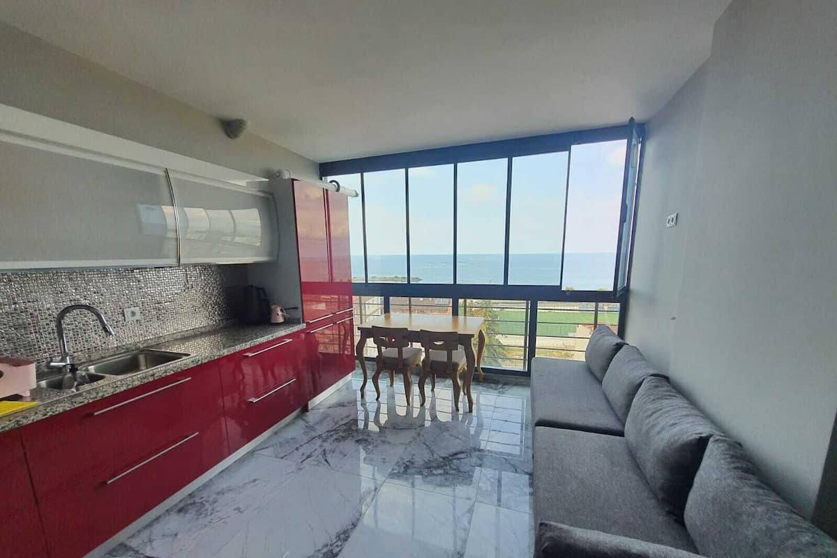 A modern kitchen with a marble floor, red cabinets, and a view of the sea through large windows.