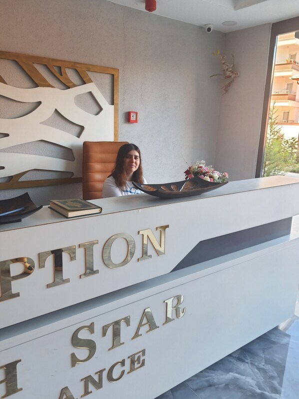 A reception desk with a woman seated behind it, a decorative wall panel, and a window with a view of buildings. - North Star Apart (Yomra)