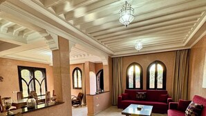 A spacious living room with a red sofa, a coffee table, and a chandelier. There are two arched windows with curtains, a wooden chair, and a small table with a glass container. - Vacation Riad with private pool 3 CH Hivernage (Marrakech)