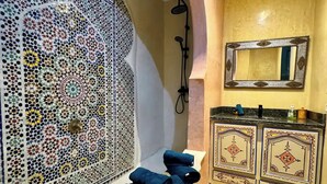 A bathroom with a decorative tiled wall, a shower, a sink with a mirror, and towels on a bench. - Vacation Riad with private pool 3 CH Hivernage (Marrakech)