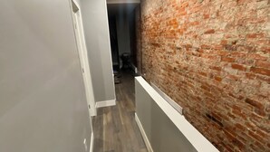 Un couloir avec un mur de briques d’un côté et un mur blanc de l’autre, qui mène à une pièce au plancher de bois.