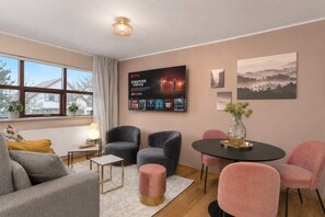 A modern living room with a sofa, two armchairs, a coffee table, and a dining table. There is a large poster on the wall and a window with curtains.
