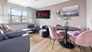 Ett modernt vardagsrum med en grå soffa, ett runt bord och rosa stolar. En platt-TV visar Netflix-logotypen.