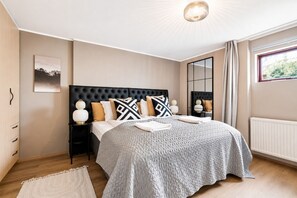 A modern bedroom with a tufted headboard, a large bed with patterned bedding, bedside lamps, a view of greenery through a window, and a radiator. - Inviting 2BR Haven in the Heart of the Suburbs (Reykjavík)