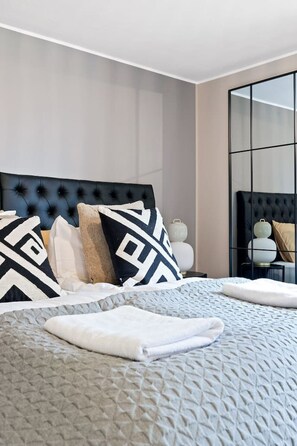 Une chambre à coucher moderne avec une tête de lit capitonnée, un grand lit agrémenté d’oreillers à motifs, une table de chevet avec une lampe et un grand miroir qui reflète la pièce.