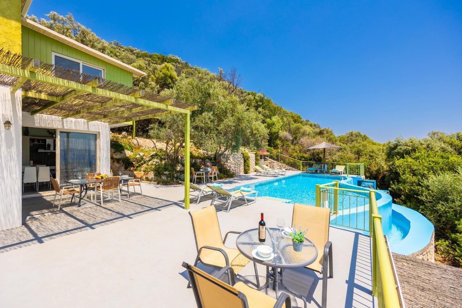 Uma área de piscina com espreguiçadeiras, uma mesa com garrafa e copo, e um pérgula.