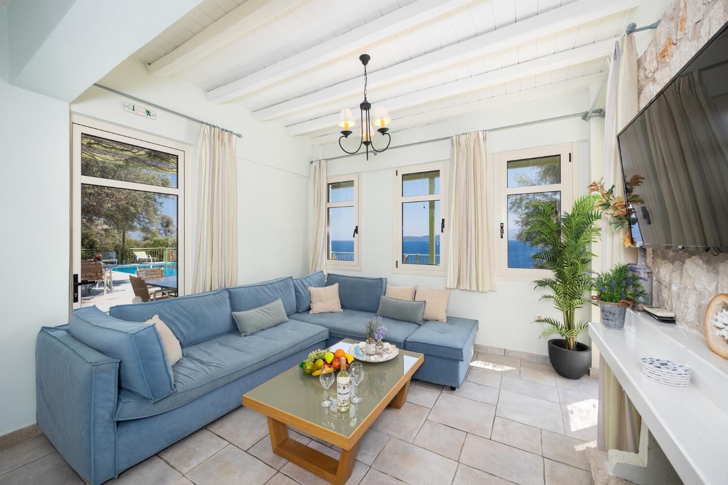 A spacious living area with a blue sofa, a coffee table with fruits and glasses, and large windows offering a view of the sea.