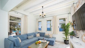 A spacious living area with a blue sofa, a coffee table with fruits and glasses, and large windows offering a view of the sea.