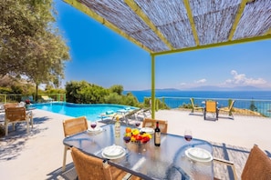 Zona de comedor junto a la piscina con una mesa preparada para dos, con vistas a un mar azul cristalino y un paisaje montañoso.