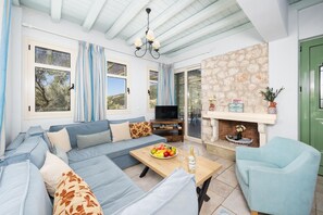 A living room with a sofa, a television, a fireplace, and a dining table with fruit and wine.