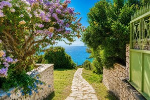 A stone pathway leading to a view of the sea, flanked by lush greenery and flowering plants.