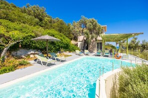 A swimming pool with lounge chairs and a pergola, surrounded by greenery and a stone building.
