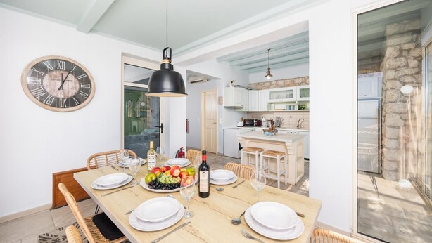 A dining area with a wooden table set for four, a clock on the wall, and a kitchen in the background. - Villa Anemus - Three Bedroom Villa, Sleeps 6 (Lefkada)