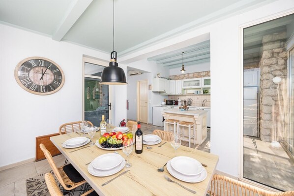 A dining area with a wooden table set for four, a clock on the wall, and a kitchen in the background.