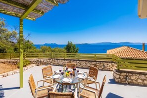 An outdoor dining area with a table set for four, surrounded by wicker chairs, overlooking a scenic view of the sea and mountains. - Villa Anemus - Three Bedroom Villa, Sleeps 6 (Lefkada)