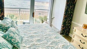 Un dormitorio con balcón que da al mar, una cama con ropa de cama estampada y un aparador con cajones.