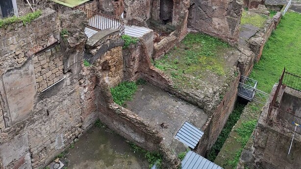 An ancient ruin with stone walls and a small courtyard, surrounded by modern buildings.