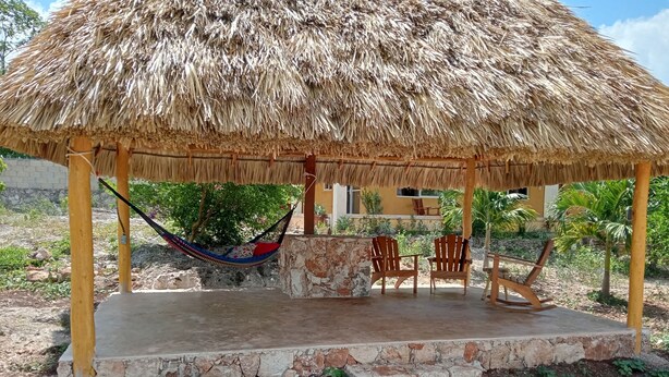 A thatched-roof gazebo with a hammock, wooden chairs, and a stone bench.