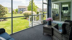 Un balcón con vistas a un césped verde y casas. Hay una silla de mimbre y una mesita con un jarrón de flores.