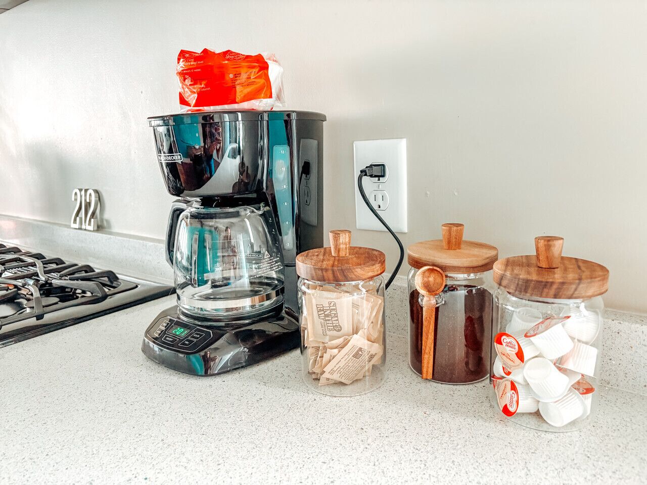 A kitchen counter with a coffee maker, two jars with wooden lids, and a bag of sugar.