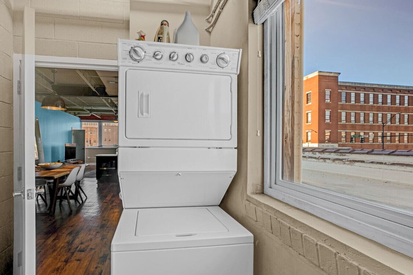A compact kitchen with a built-in washer and dryer, a dining area with a table and chairs, and a view of a brick building through a window.