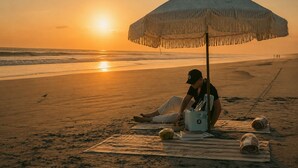 Escena de playa con una persona sentada bajo un paraguas a rayas, un refrigerador y una toalla enrollada sobre la arena.