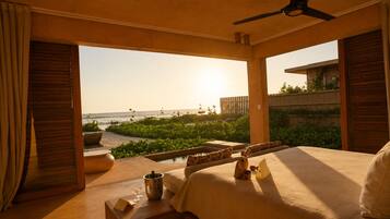 A hotel room with a bed, a small table with a bottle and glasses, a view of the ocean, and a ceiling fan.