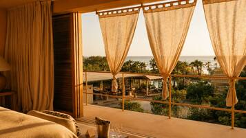 A hotel room with a large window, a bed, a bedside table with a lamp, and a view of the ocean and resort area.