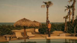 Un hombre está de pie junto a una piscina con sillas de descanso y palmeras.
